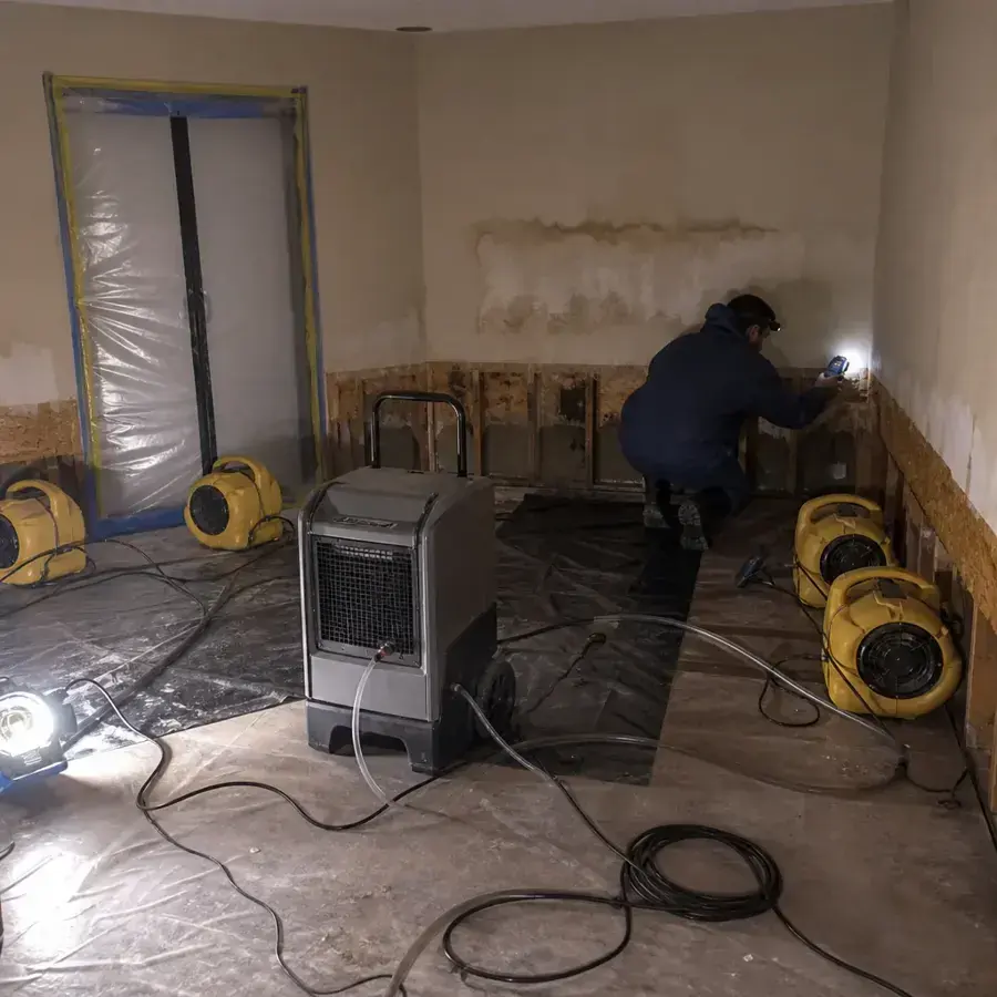 Technician checking a flood-cut wall beside a dehumidifier and air movers under plastic containment